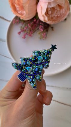 a hand holding a blue beaded christmas tree brooch in front of pink roses