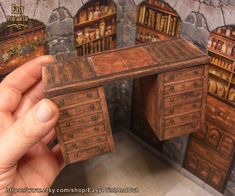 a hand holding a miniature wooden desk in front of a wall with bookshelves