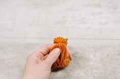 a hand holding an orange piece of yarn on top of a cement floor next to a person's hand