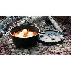 an open pot with eggs and carrots in it on the ground next to a campfire