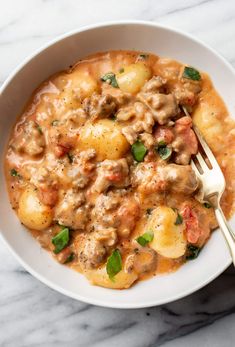 a white bowl filled with meat and potatoes on top of a marble table next to a fork