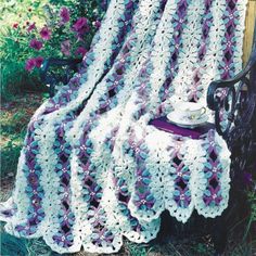 a crocheted blanket sitting on top of a wooden bench next to purple flowers