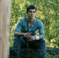 a young man sitting on top of a log in the woods with his hands folded