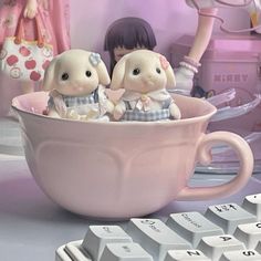 two little dolls are sitting in a pink cup next to a computer keyboard and mouse