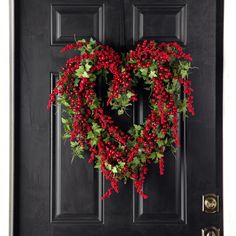 a heart - shaped wreath hangs on the front door of a black door with red berries and green leaves