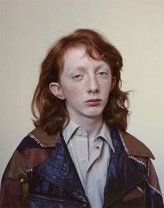 a woman with red hair wearing a leather jacket and looking at the camera while standing in front of a white wall