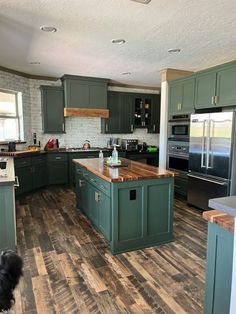 a kitchen with green cabinets and wood flooring, an island in the middle is surrounded by stainless steel appliances