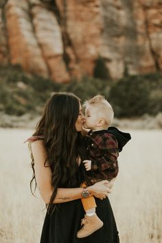 a woman holding a baby in her arms and kissing it's face with mountains in the background