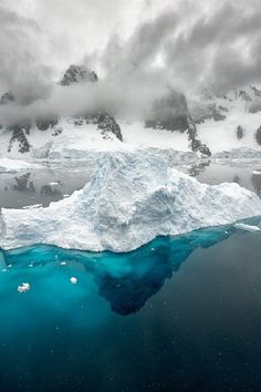 an iceberg floating in the ocean with mountains in the background and cloudy skies above