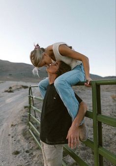 a man and woman kissing on the side of a road near a green fence with mountains in the background