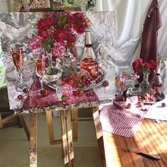 an easel with flowers and wine glasses on it in front of a painting that is being displayed