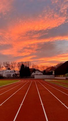 the sun is setting over a running track with buildings in the background and trees on either side