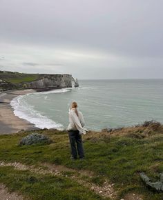 a woman standing on top of a lush green hillside next to the ocean and cliffs