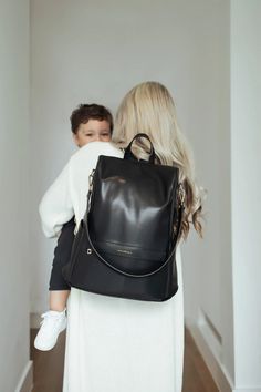 a woman carrying a baby in a black leather backpack while walking down a hall way