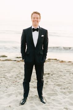 a man in a tuxedo standing on the beach with his hands in his pockets