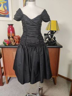 This is a beautiful show stopping well made 80s cocktail dress by an English label that I think says Carmonia Collection in a size 12uk The fabric is a swishy black tafetta like fabric ( actually polyester) and  fully lined. The dress has a wide neckline that sits on the edge of the shoulders with little pleated cap sleeves and a fitted rouched  bodice and a dropped waist. The skirt is gathered with a tuile underskirt between the outer and the acetate lining. No prickles.  The condition is very Fitted Party Dress With Gathered Skirt, Fitted Dress With Gathered Skirt For Party, Black Party Dress With Gathered Skirt, Fitted Black Dress With Gathered Skirt, Black Fitted Dress With Gathered Skirt, Fitted Midi Dress With Gathered Skirt For Party, Cocktail Dresses With Gathered Waist And Fitted Style, Vintage Ruffled Midi Dress For Evening, Formal Dress With Gathered Skirt And Fitted Bodice