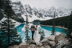 a bride and groom standing on the edge of a cliff overlooking a lake surrounded by mountains