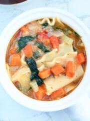 a bowl of soup with carrots, spinach and pasta in it on a marble table