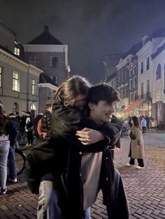 two people hugging each other in the middle of a city street at night with lots of people walking around