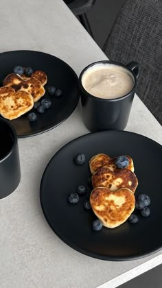 two black plates topped with pancakes and blueberries next to a cup of coffee on a table