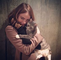 a woman holding a cat in her arms while standing next to a wall and looking at the camera