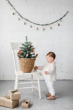 a baby standing next to a white chair with a christmas tree in the basket on it