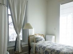 a bed sitting next to a window in a room with white curtains on the windowsill