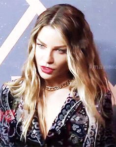 a close up of a person with long hair wearing a shirt and necklace, standing in front of a wall