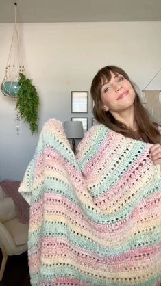 a woman holding up a multicolored crocheted blanket in her living room