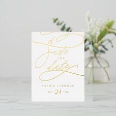 a white and gold save the date card next to a vase with flowers in it