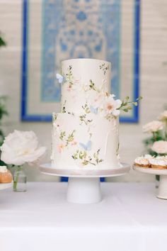 a white wedding cake with flowers on the top is sitting on a table next to cupcakes