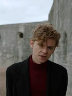 a young man with curly hair standing in front of a stone wall and looking at the camera