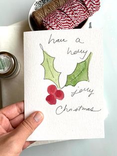 a hand holding a christmas card with holly on it and some twine spools