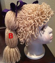 a white mannequin head with purple ribbon on it and two balls of yarn in the hair