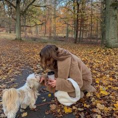 a woman kneeling down next to a white dog on top of leaf covered ground with trees in the background