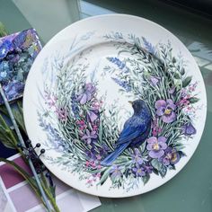 a blue bird sitting on top of a white plate with purple flowers and greenery