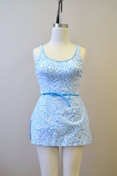 "Description: 1960s blue and white floral printed skirted swimsuit. 65% polyester, 35% cotton. Back zipper and hook and eye closures, and it also has some button interior closures for a snug fit. It has a soft cup bra and a tie belt. The sides have smocked panels, and the bodice has some boning. The skirt has side vent slits. Condition: In great condition, with no holes. There is some yellowing inside the bra cups, but it is not visible when being worn. Designer: Perfection Fit by Roxanne Stated Spring Fitted Lined Tankini, Fitted Lined Tankini For Spring, Fitted Blue Swim Skirt With Built-in Bra, Fitted Blue Swim Skirt For Spring, Sleeveless Cotton Swimwear, Retro Blue Lined Swimwear, Vintage Blue Swimwear For Summer, Spring Fitted Blue Swim Dress, Blue Fitted Swim Dress For Spring