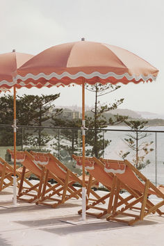 A serene outdoor patio setup with coral-colored beach umbrellas featuring scalloped edges and matching deck chairs. The view overlooks a coastline lined with trees and distant homes, creating a coastal retreat ambiance. Beach Cart, Picnic Set