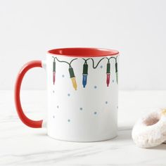 a red and white coffee mug with christmas lights on it next to a donut