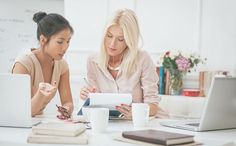 two women sitting at a table with laptops and papers in front of them, one writing on a piece of paper