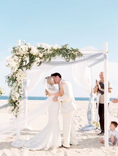 a bride and groom kiss under an arch on the beach as their guests take pictures