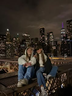 two young women sitting on a bridge in front of the city skyline at night time