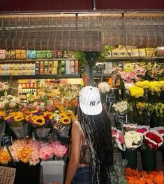 a woman with long black hair standing in front of a flower shop filled with lots of flowers