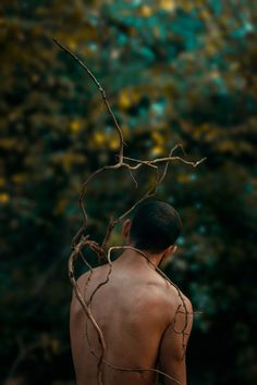 a man with no shirt on standing in front of some trees and bushes, looking down at the ground