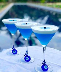 three martini glasses with blue liquid and an evil eye on the rim are sitting on a white table cloth