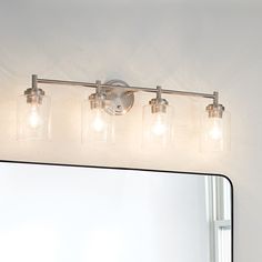 a bathroom vanity light with four lights on it and a mirror above it, in front of a white wall