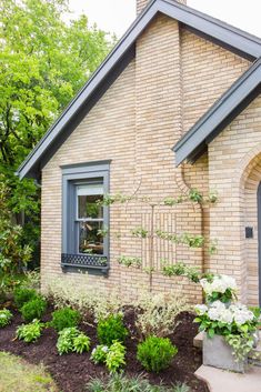 a brick house with blue shutters and flowers in the front yard on a sunny day