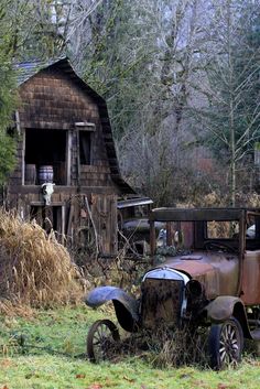 an old rusted out truck sitting in front of a wooden building with a barn behind it