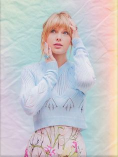 a woman with blonde hair wearing a blue sweater and floral print skirt holding her hand to her ear