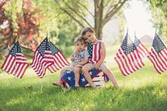 two young children sitting on top of american flags in the grass with trees behind them